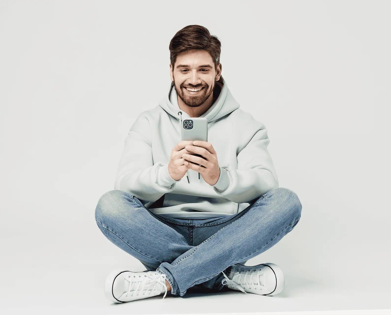 Smiling young man in a hoodie sitting cross-legged on the floor and using a smartphone.