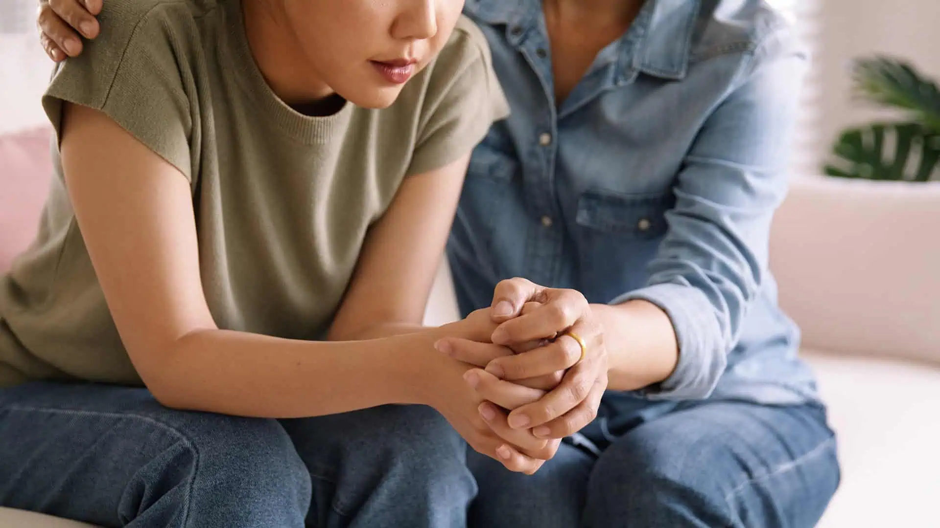 Close-up of a woman being comforted by another person, hands clasped and arms around her.