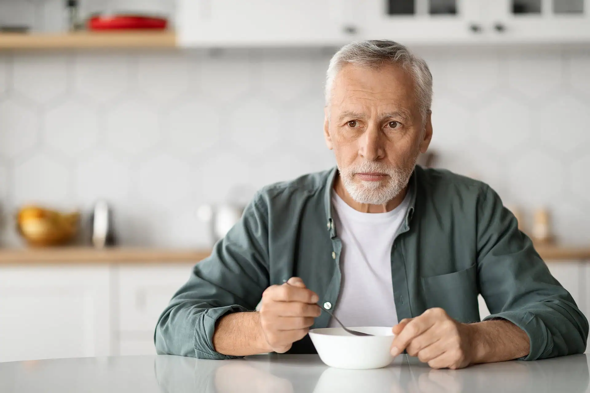 Older man with grey hair and beard sitting at a table, holding a spoon over a bowl of soup while looking off to the side.