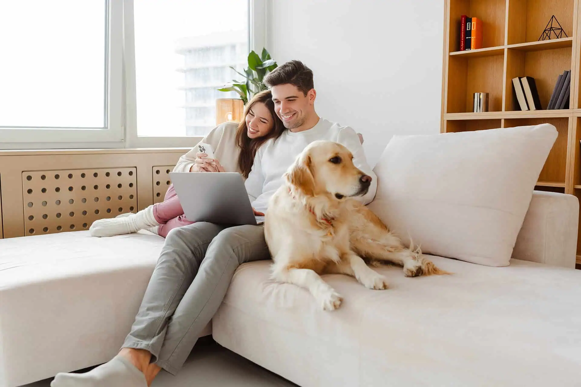 Smiling couple sitting on a sofa with a golden retriever by their side, using a laptop together.