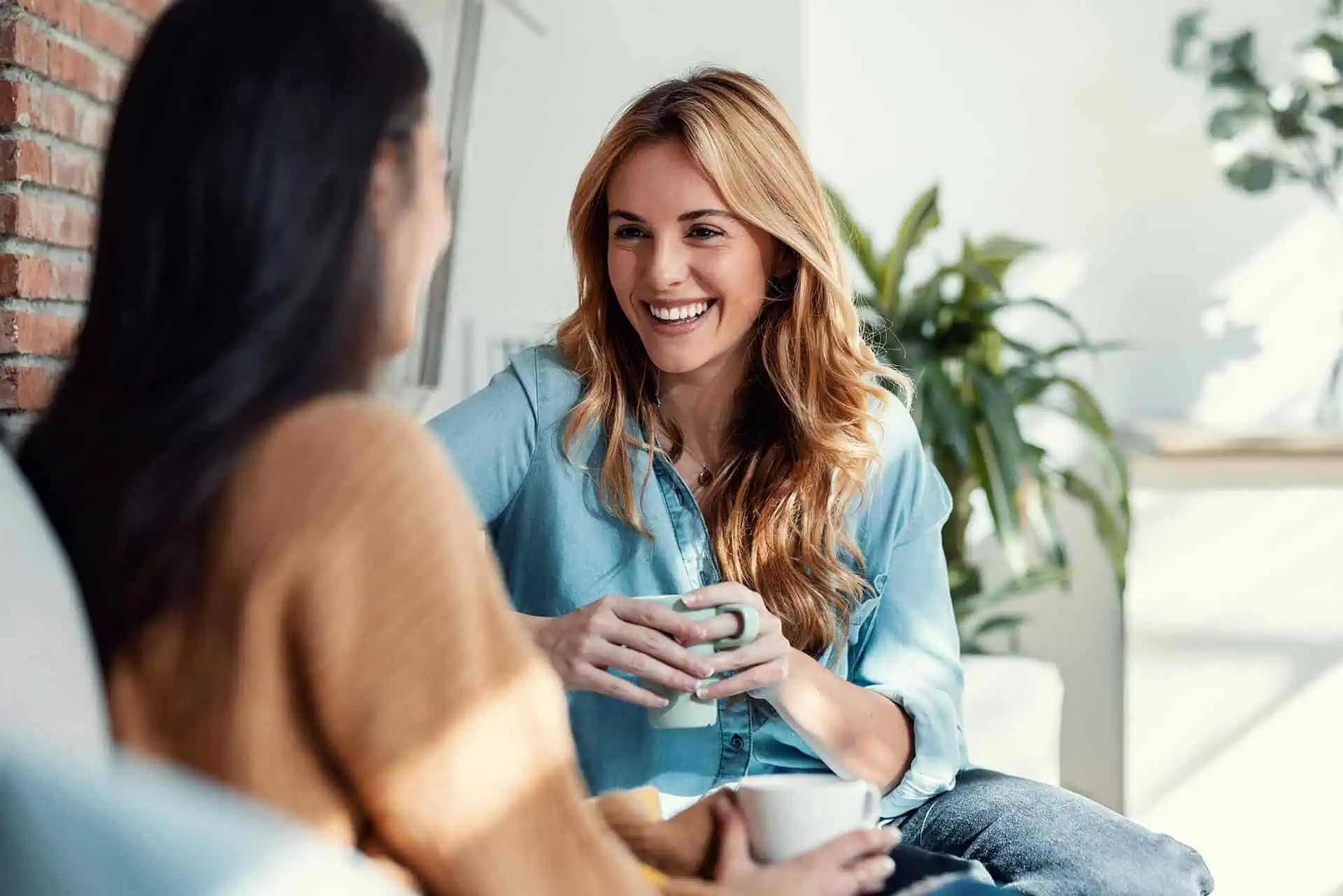 Two women sitting and chatting while holding coffee mugs, smiling in a cozy setting.