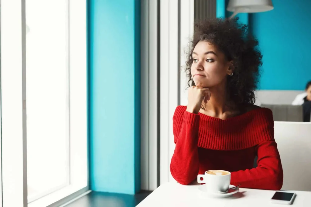 Pensive woman with curly hair in a red sweater sitting at a cafe table with a cup of coffee, looking thoughtfully out of the window.