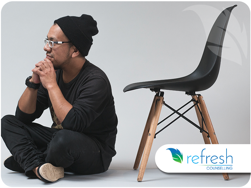 Man sitting on the floor next to an empty chair, wearing a beanie and glasses, looking contemplative.
