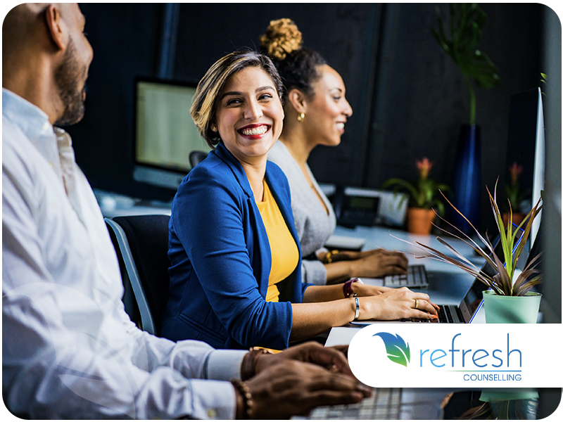 Smiling woman in a blue blazer seated at a table with coworkers, working on a computer.