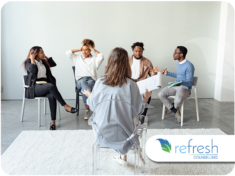 Group of five adults in chairs during a group therapy session, some gesturing and one woman covering her ears, with a facilitator holding a clipboard facing them.