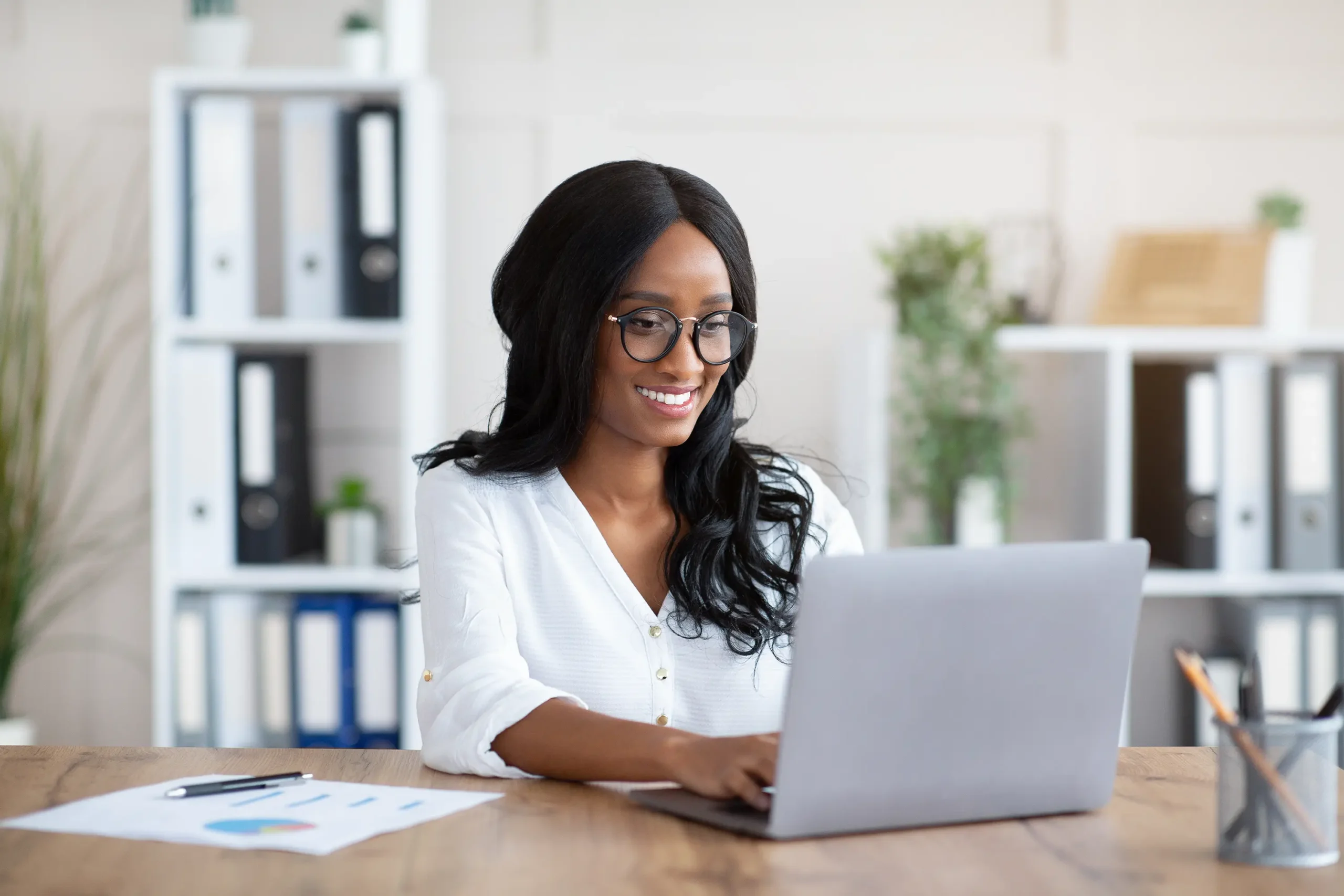 Smiling woman with glasses working on a laptop at her desk in an office.