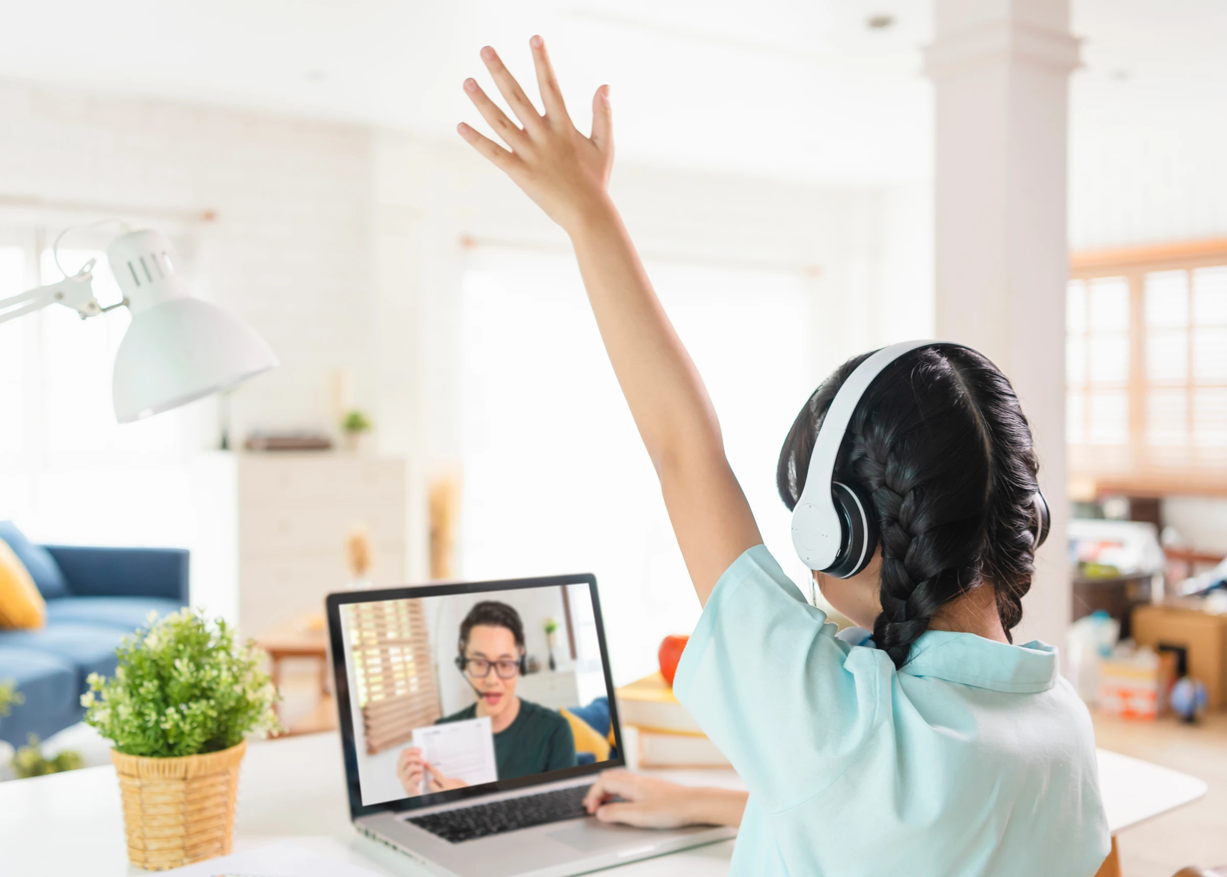 Young girl wearing headphones raising her hand while attending an online class on a laptop.
