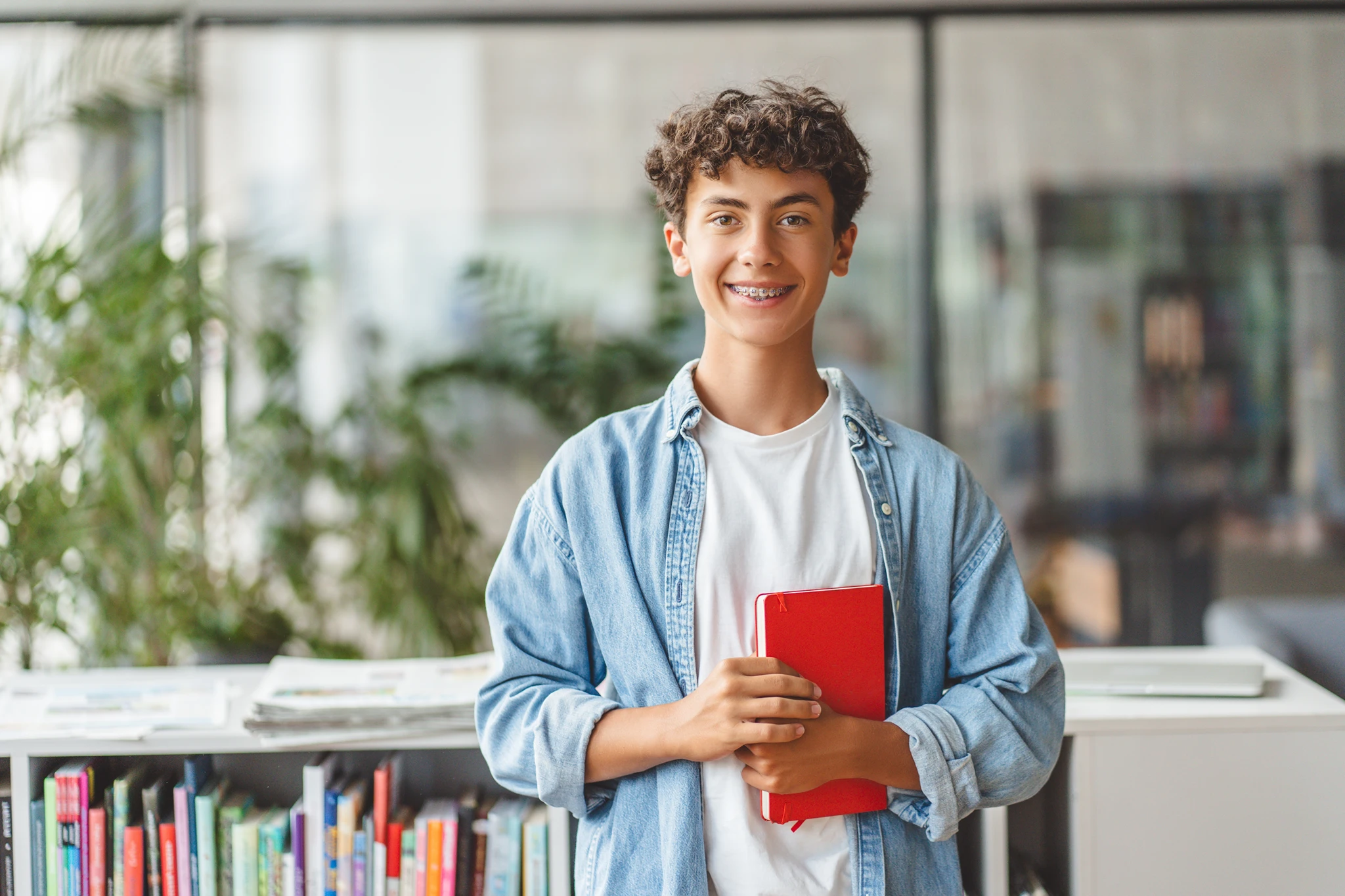 Smiling young man with curly hair holding a red notebook in a library.