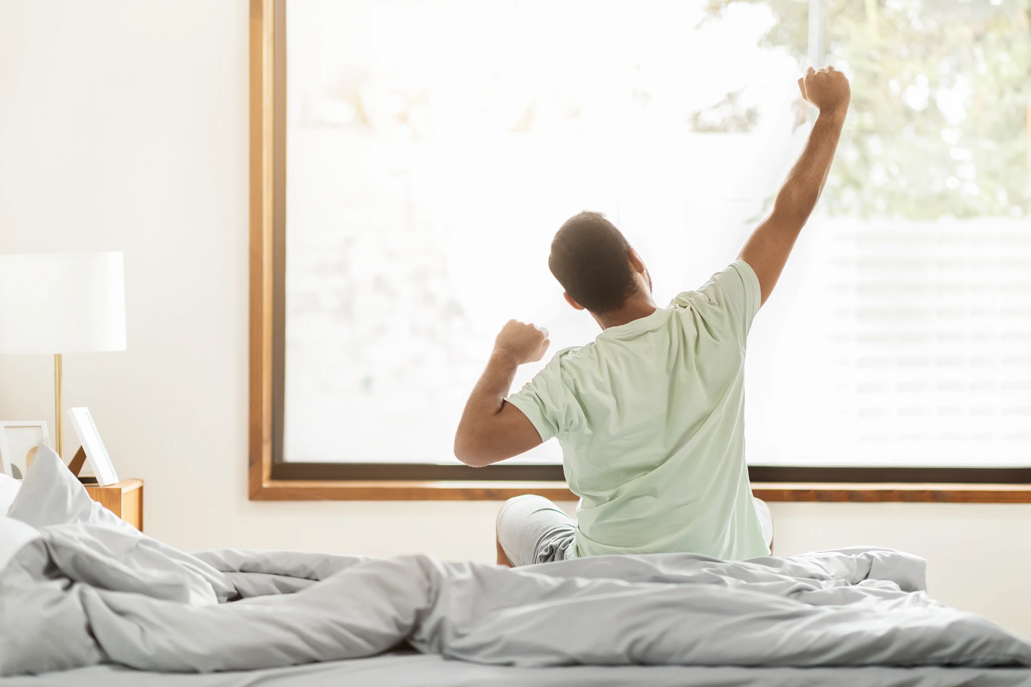 Man stretching in bed with arms raised in a bright bedroom, viewed from behind.
