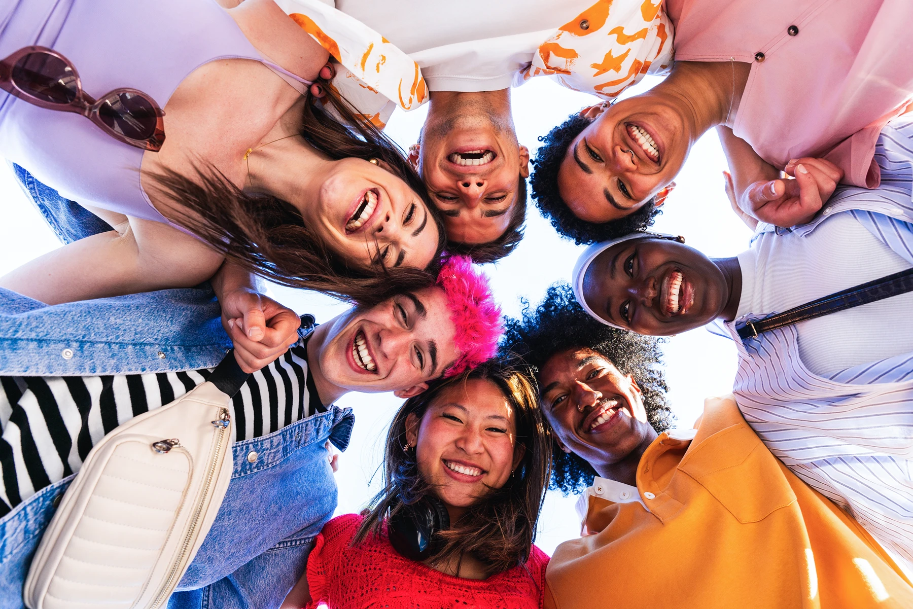 Group of diverse young friends in colorful clothes huddled in a circle, smiling and looking down at the camera.