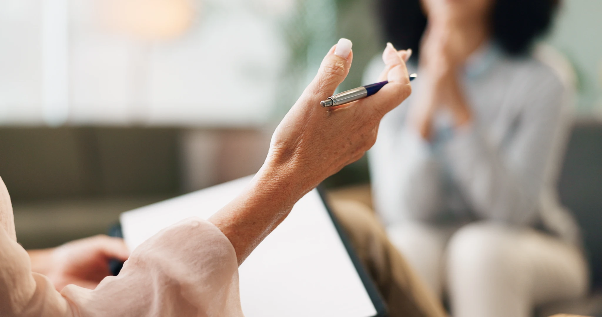 Close-up of hands holding a pen and clipboard during a conversation, with a person in the background.