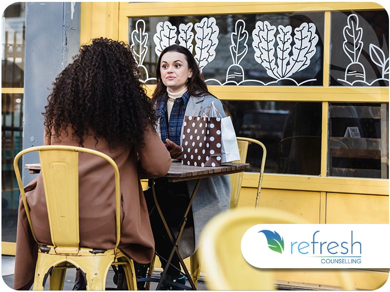 Two women talking over coffee at an outdoor café table, one with her back to the camera.