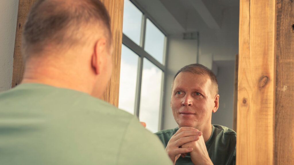 Image of a man looking at his reflection in a mirror, standing with hands clasped, in a room with large windows.