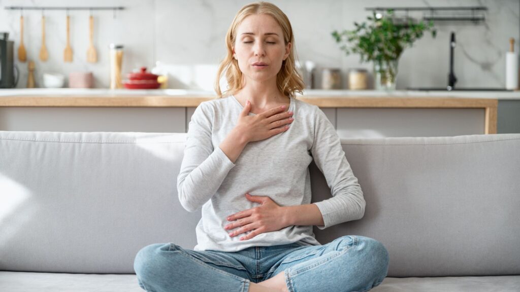 Woman meditating on a couch with hands on chest and stomach, promoting relaxation in a cozy kitchen setting.