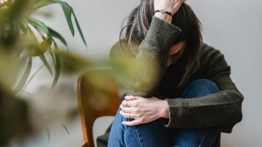 Person sitting with head in hand, showing signs of anxiety or distress, with a plant in the background.