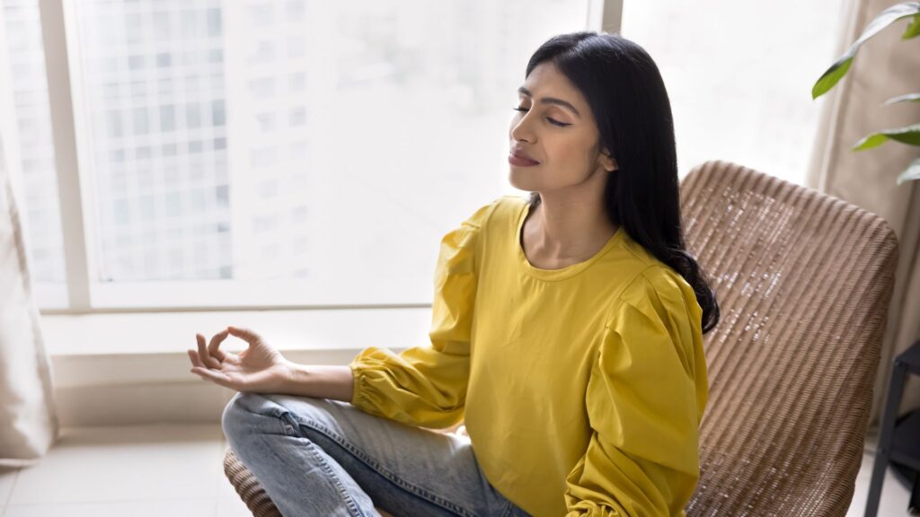 A person in a yellow top practicing meditation with hands in an "Om" gesture, sitting on a wicker chair by a window with a city view.