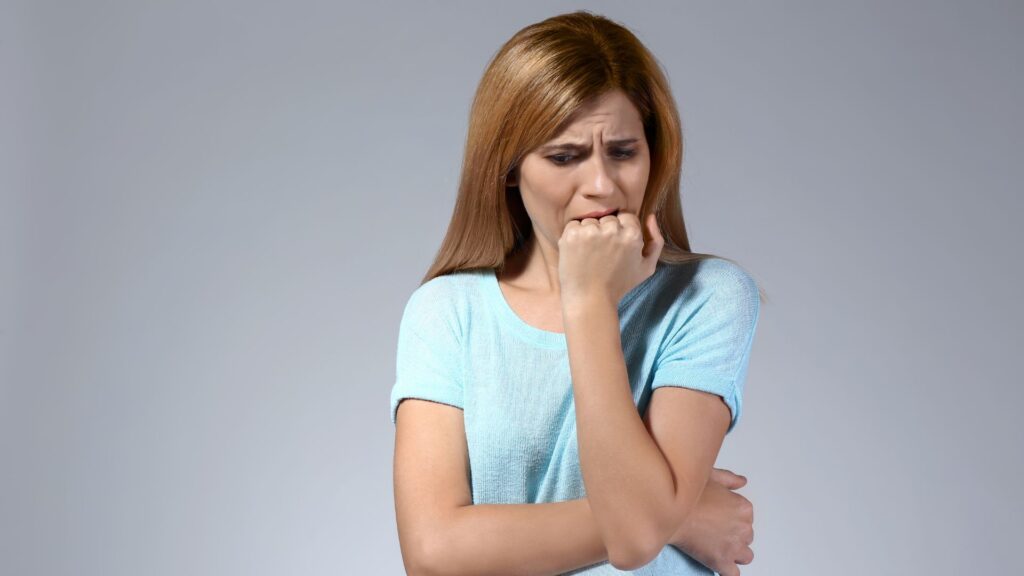 A woman in a light blue shirt biting her nails, showing anxiety against a gray background.