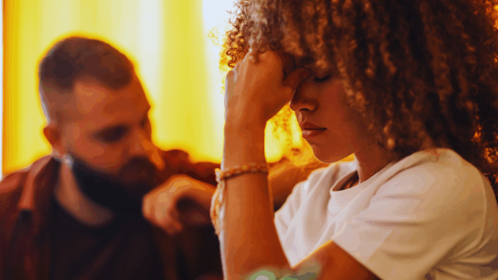 Woman with curly hair resting forehead on hand, looking distressed; man in background gazing at her. Warm lighting. Focus: emotional strain in relationships.