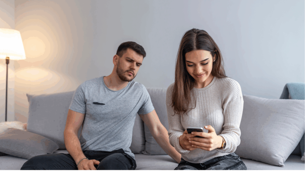 Image depicting a couple on a couch with "trust issues", showing a man looking concerned while a woman smiles at her phone, suggesting tension.