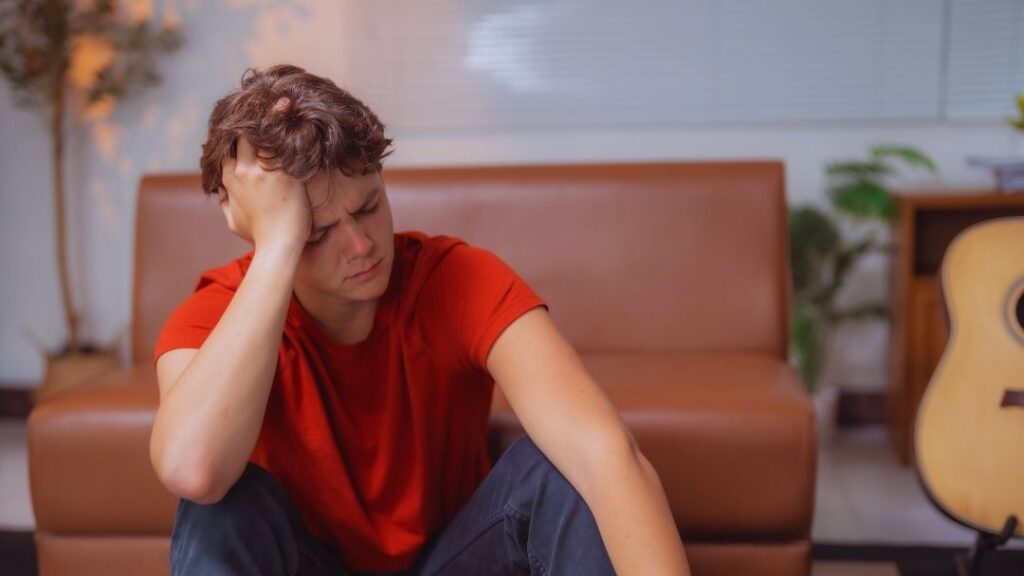 A teenage boy sitting on the floor, looking troubled and resting his head on his hand, representing a teen struggling with anxiety.