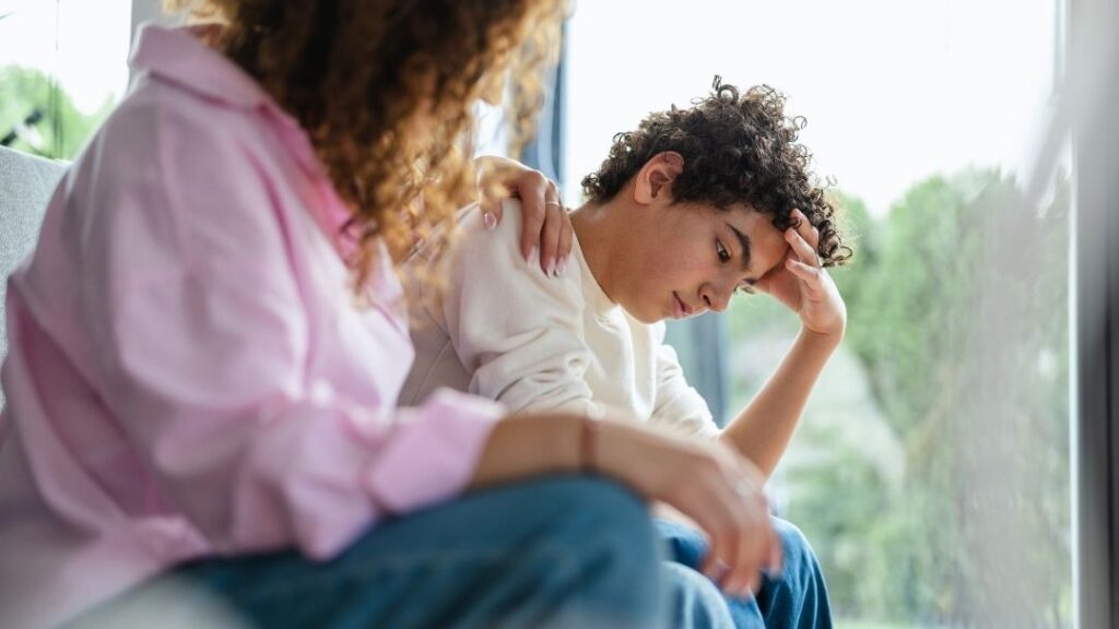 A mother comforting her teenage son who is looking down and holding his head, illustrating parental support for teen depression.