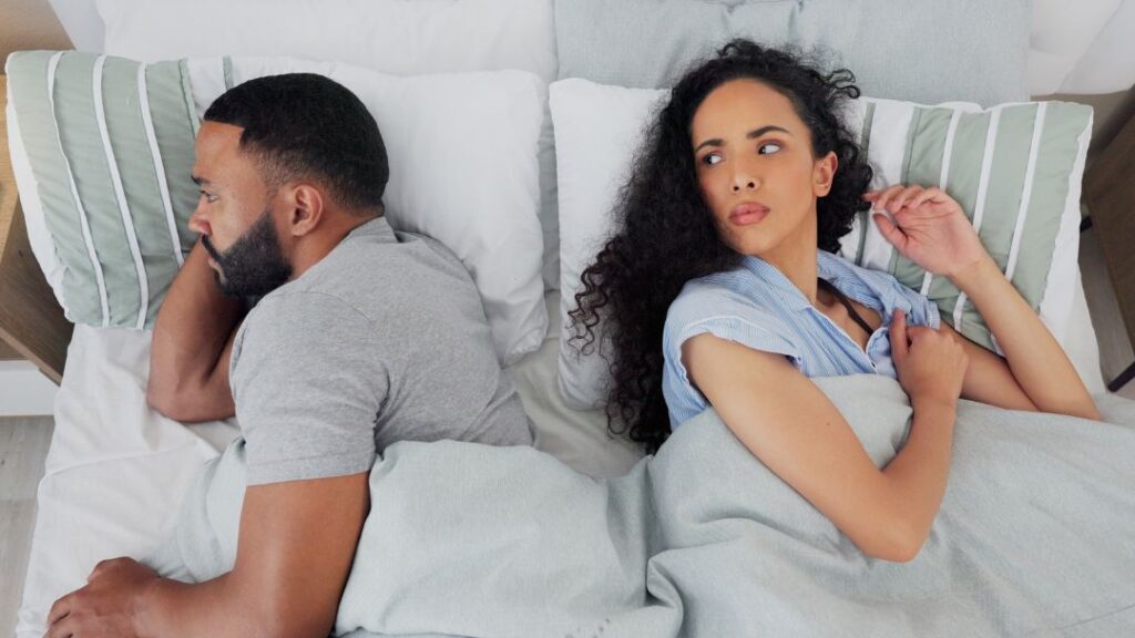 Couple lying in bed facing away from each other, showing emotional distance or disconnection.
