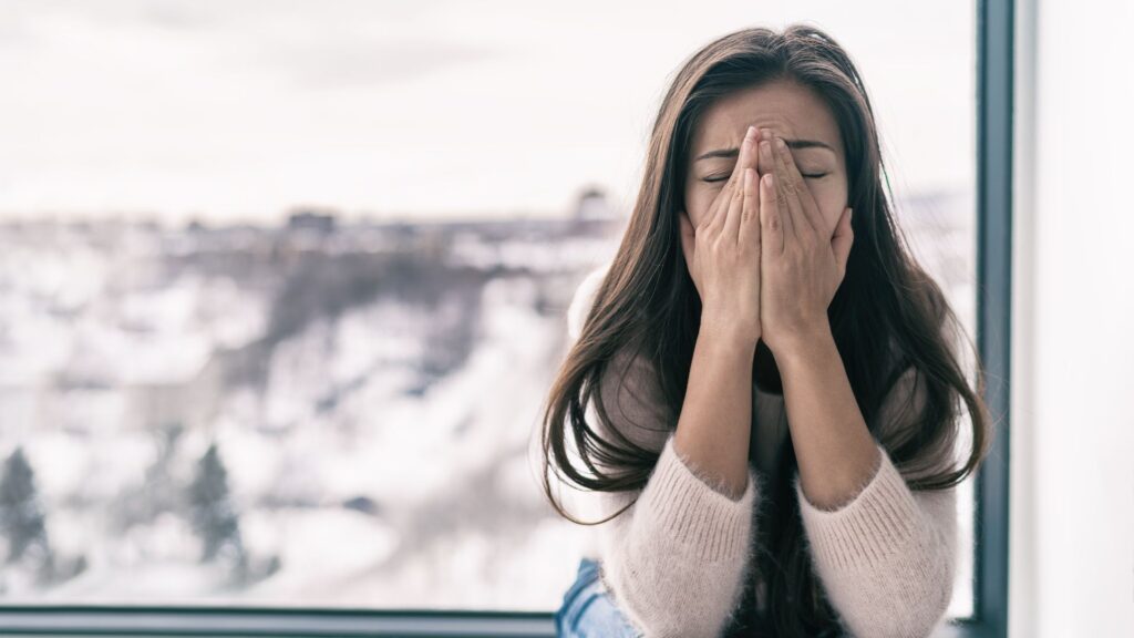 Image of a woman with hands over her face, sitting by a window with a snowy landscape, suggesting emotional distress.
