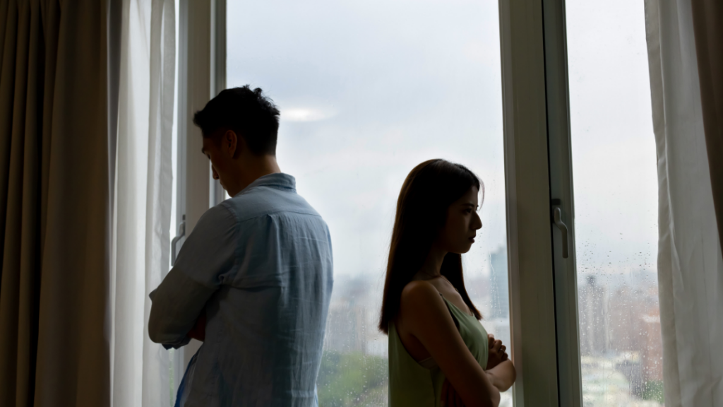 Couple experiencing relationship insecurity standing apart by window, illustrating ways to stop feeling insecure in a relationship